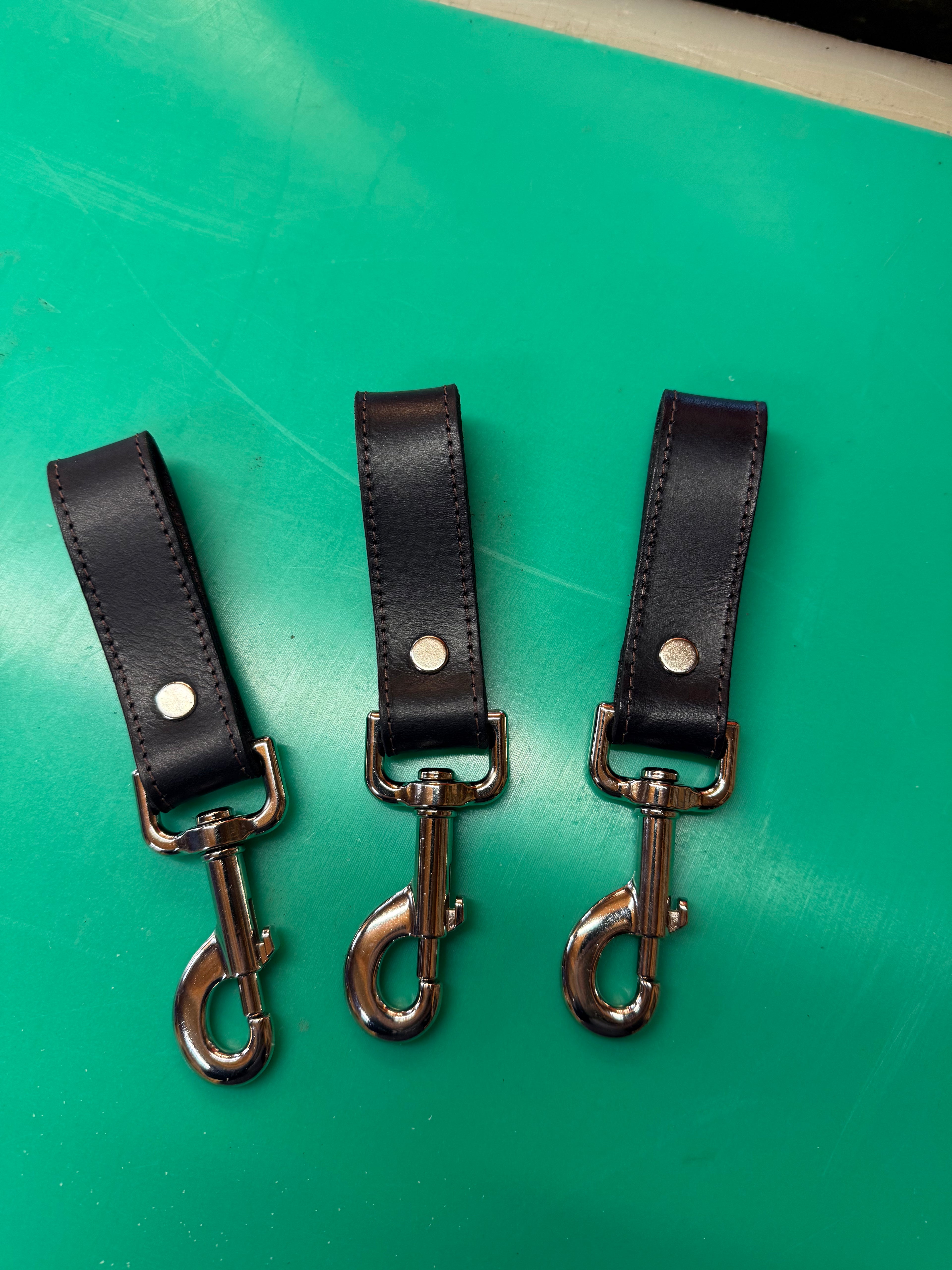 Three black leather bag clips with shiny metal snaps, arranged neatly on a green surface by FULLSUMMED FALCONRY.