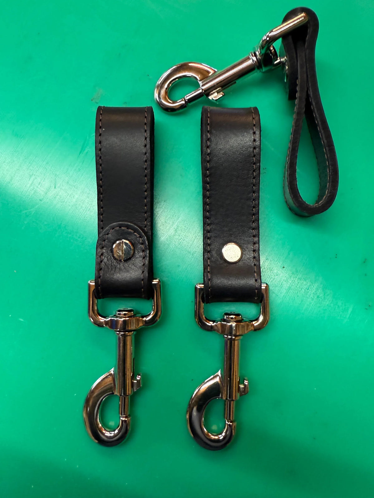 Two black leather bag clips with silver hardware on a green background, showcasing the Bag Clip by FULLSUMMED FALCONRY.